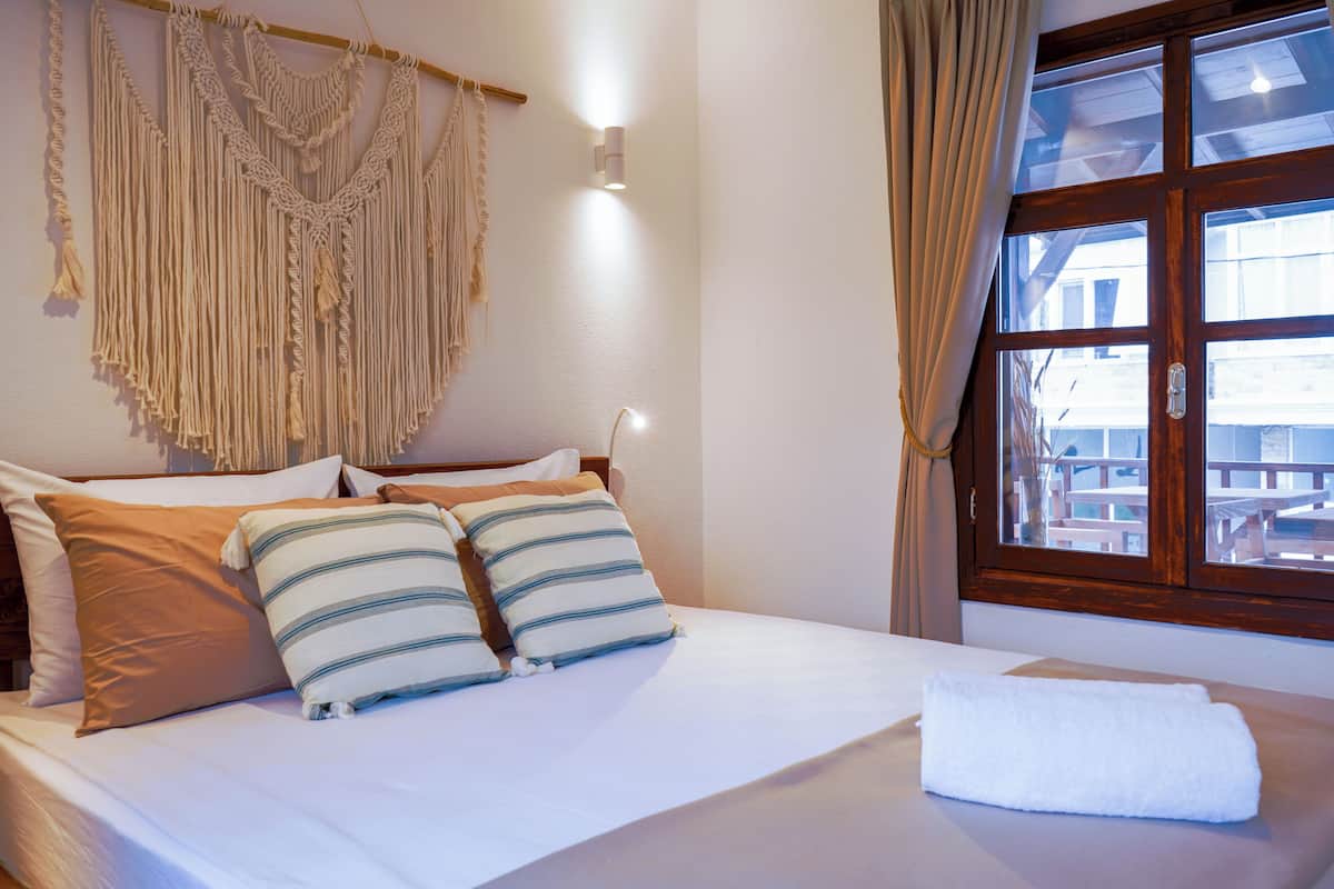 Ein ordentlich bezogenes Bett mit zwei Kissen, ein Wandbehang aus Makramee und ein Fenster mit Vorhängen.