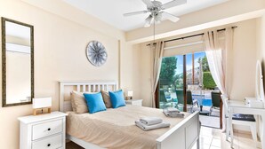 Ein Schlafzimmer mit Bett, Nachttischen, Deckenventilator, großem Fenster mit Vorhängen und Blick auf Pool und Garten.