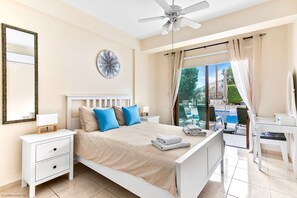 Ein Schlafzimmer mit Bett, Nachttischen, Deckenventilator, großem Fenster mit Vorhängen und Blick auf Pool und Garten.