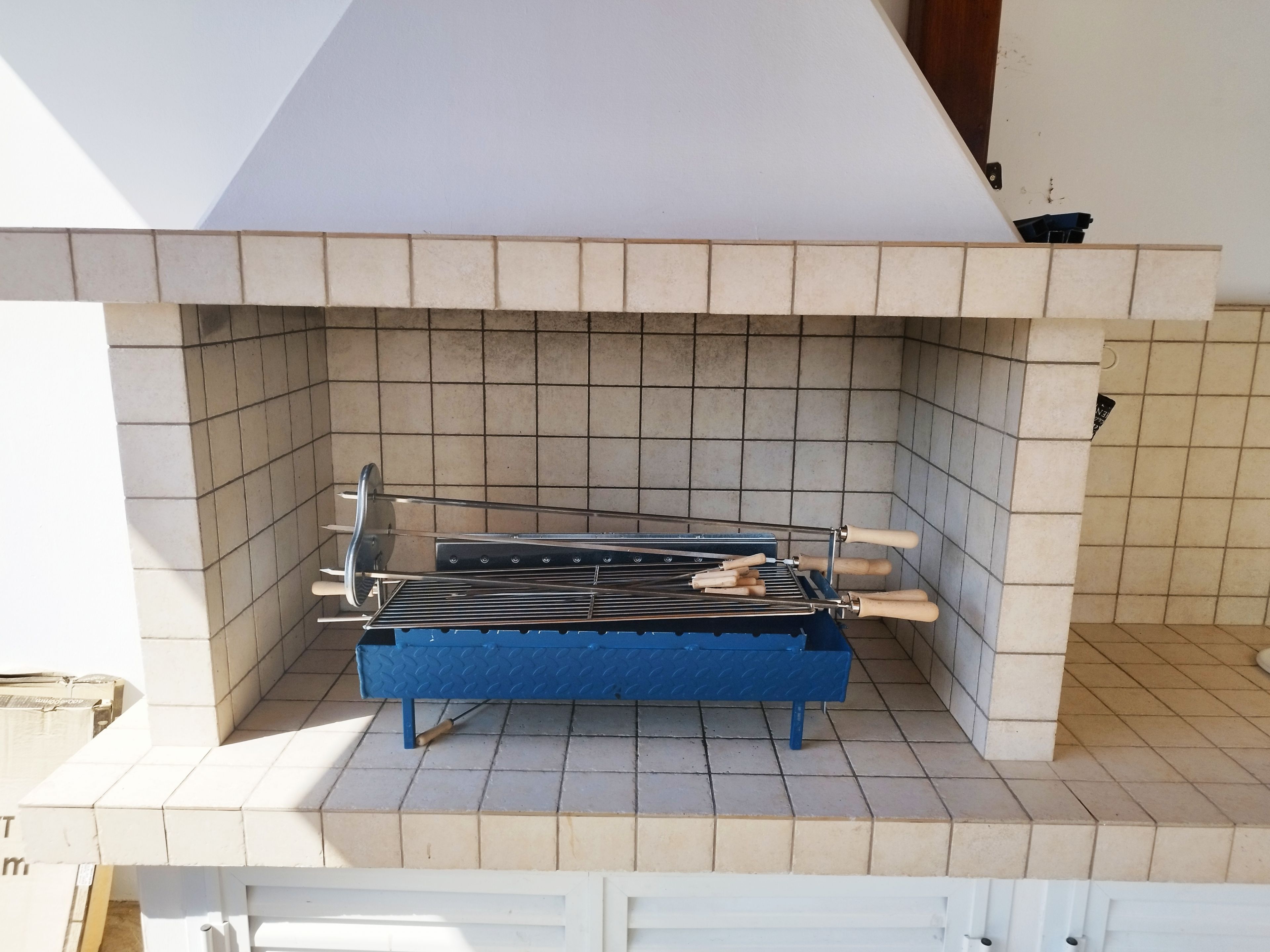 Een blauwe barbecue met een metalen rooster en houten handgrepen, op een betegelde vloer onder een wit plafond.