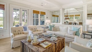A spacious living room with a large white sofa set, a wooden coffee table with decorative items, and a view of the outdoors through large windows with blinds.