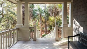 A covered porch with white railings and columns, leading to a view of a landscaped garden with palm trees and other greenery.
