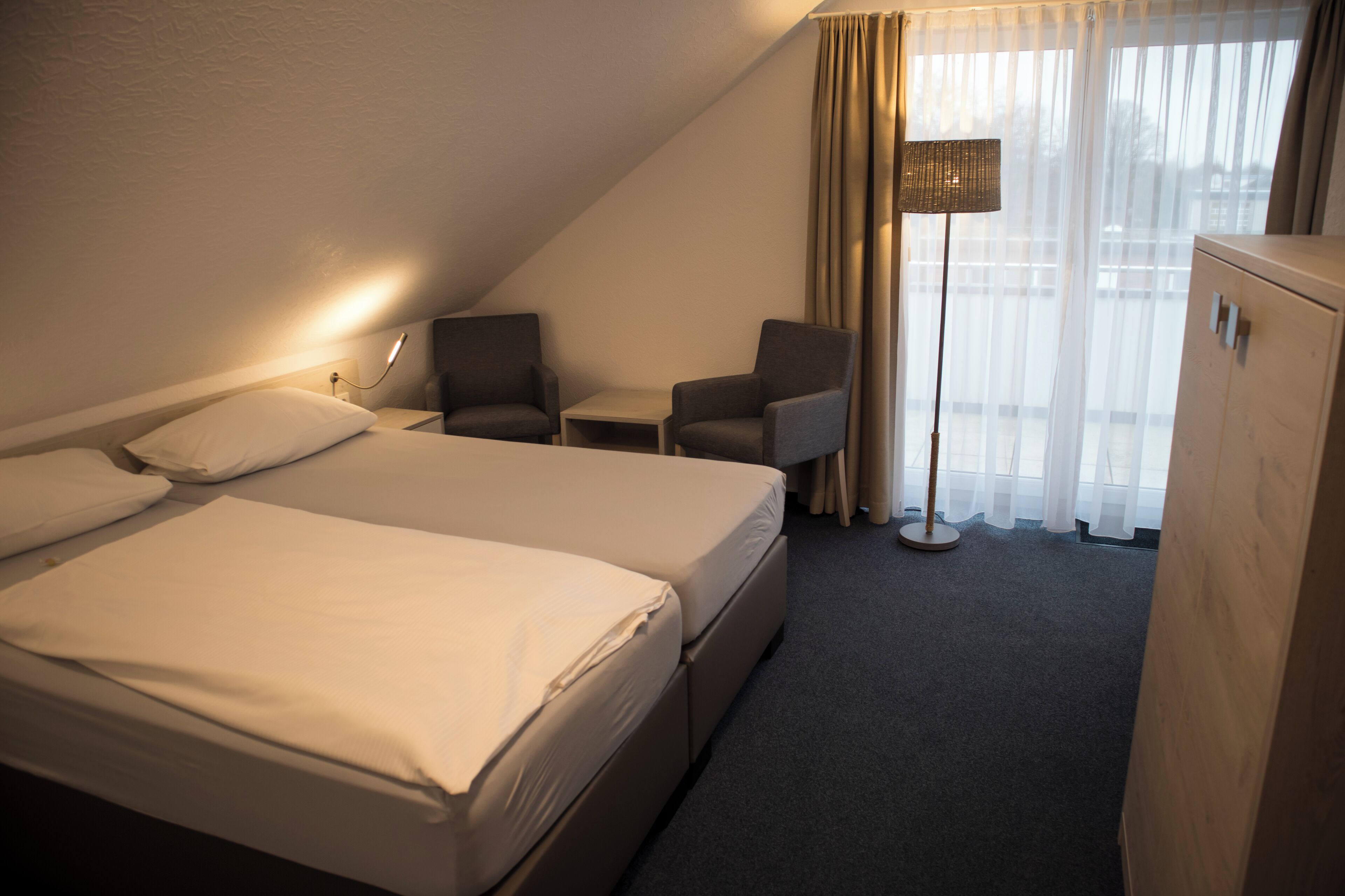 Une chambre d’hôtel avec un grand lit, deux fauteuils, une petite table, une lampe sur pied et une armoire.