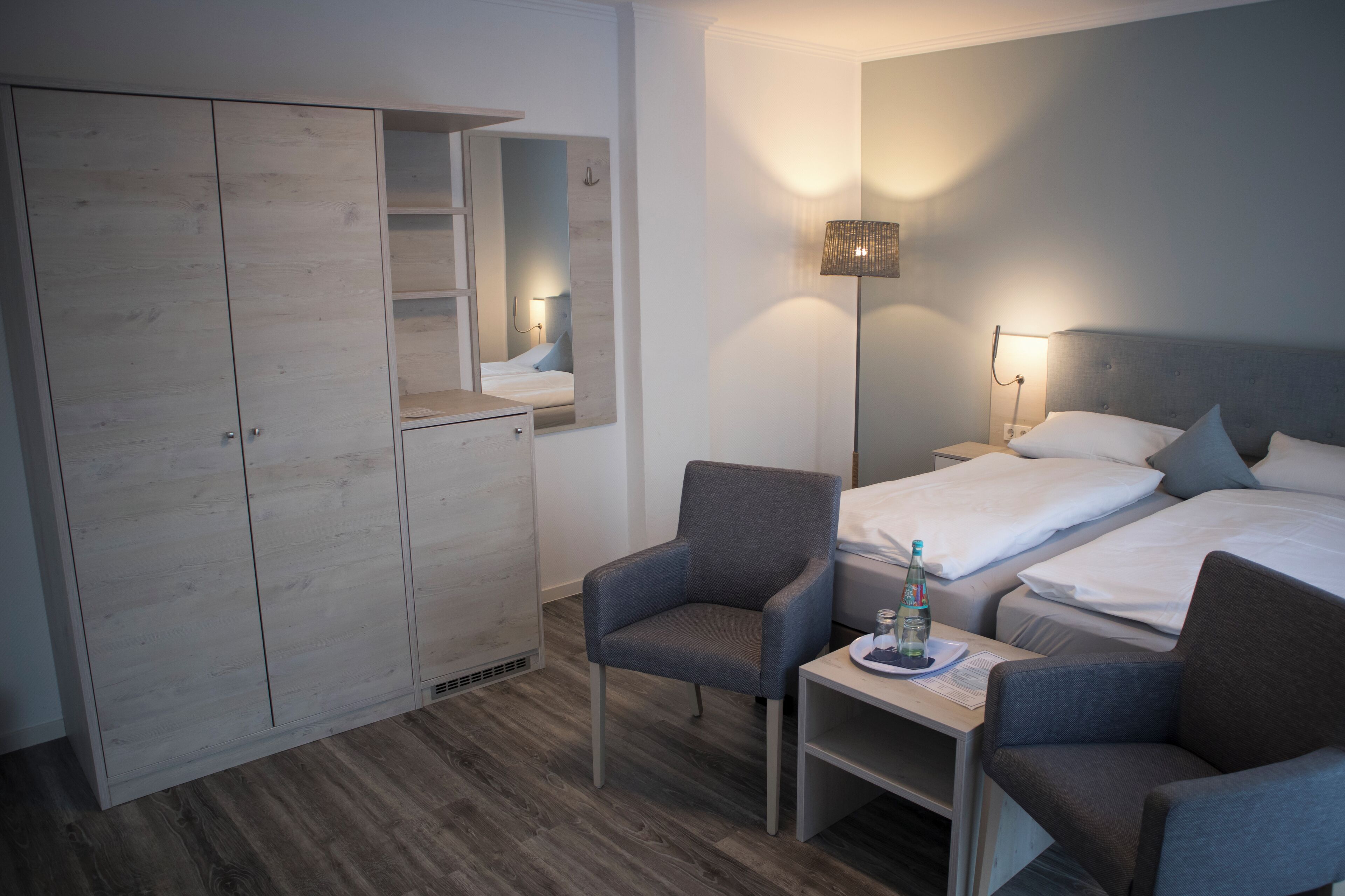 Ett hotellrum med en garderob, två enkelsängar, ett skrivbord med en stol och ett nattduksbord med en flaska och glas.
