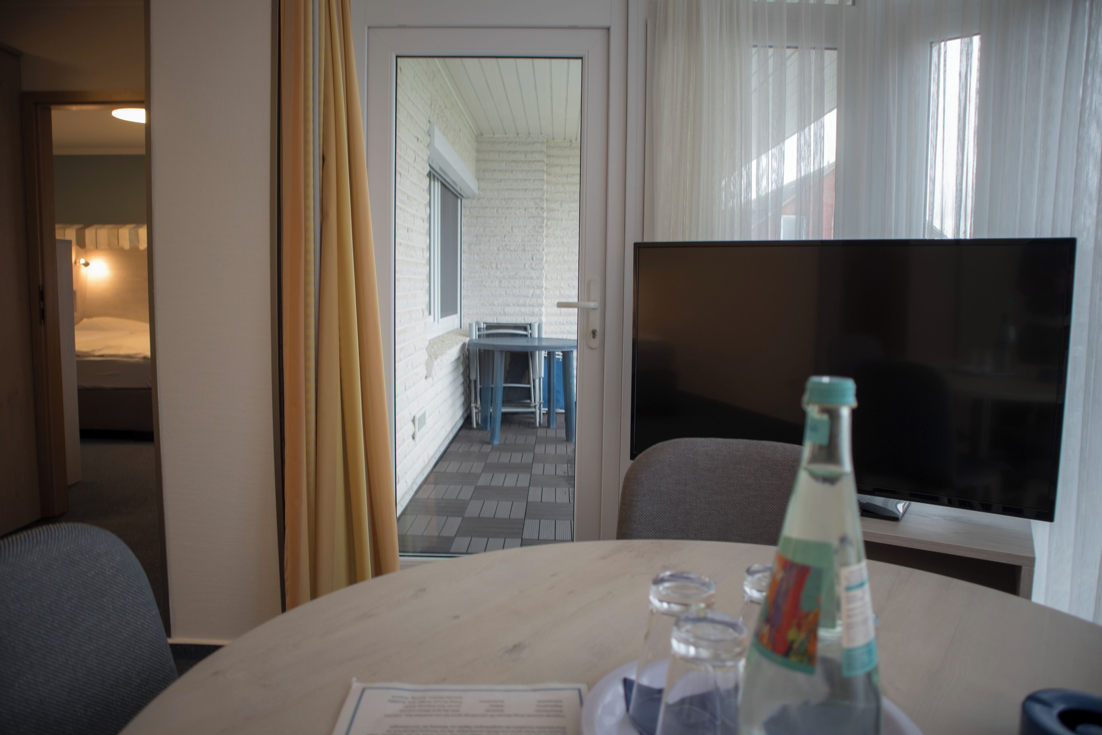 Una habitación de hotel con zona de comedor, televisión y una botella de agua sobre la mesa.
