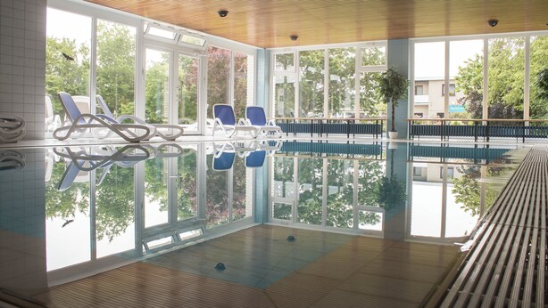 An indoor swimming pool with clear glass walls, blue lounge chairs, and a wooden ceiling.