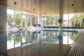 An indoor swimming pool with clear glass walls, blue lounge chairs, and a wooden ceiling. - Hotel Am Friesenstrand (Butjadingen)