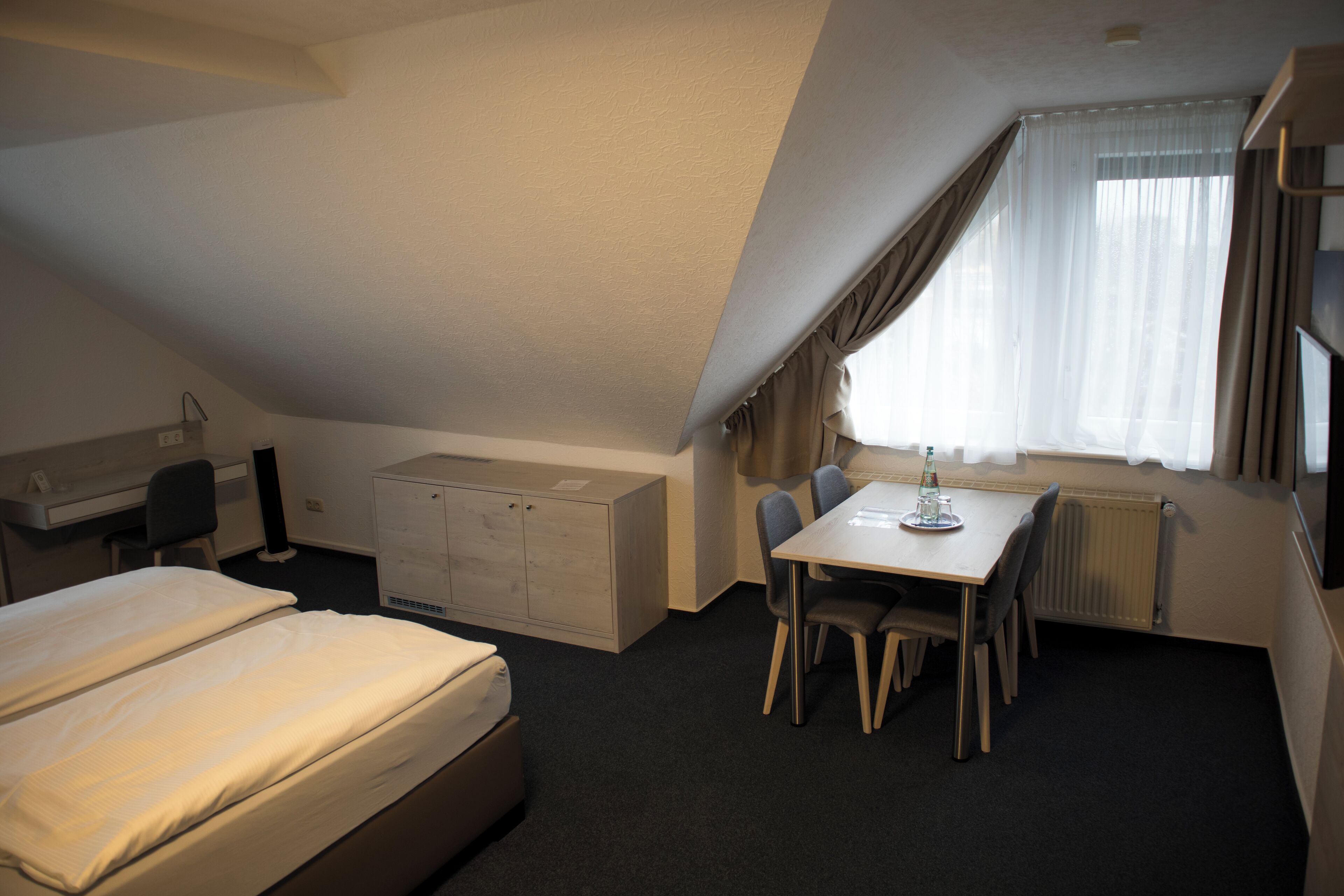 A compact hotel room with a bed, desk, chair, and a dining table.