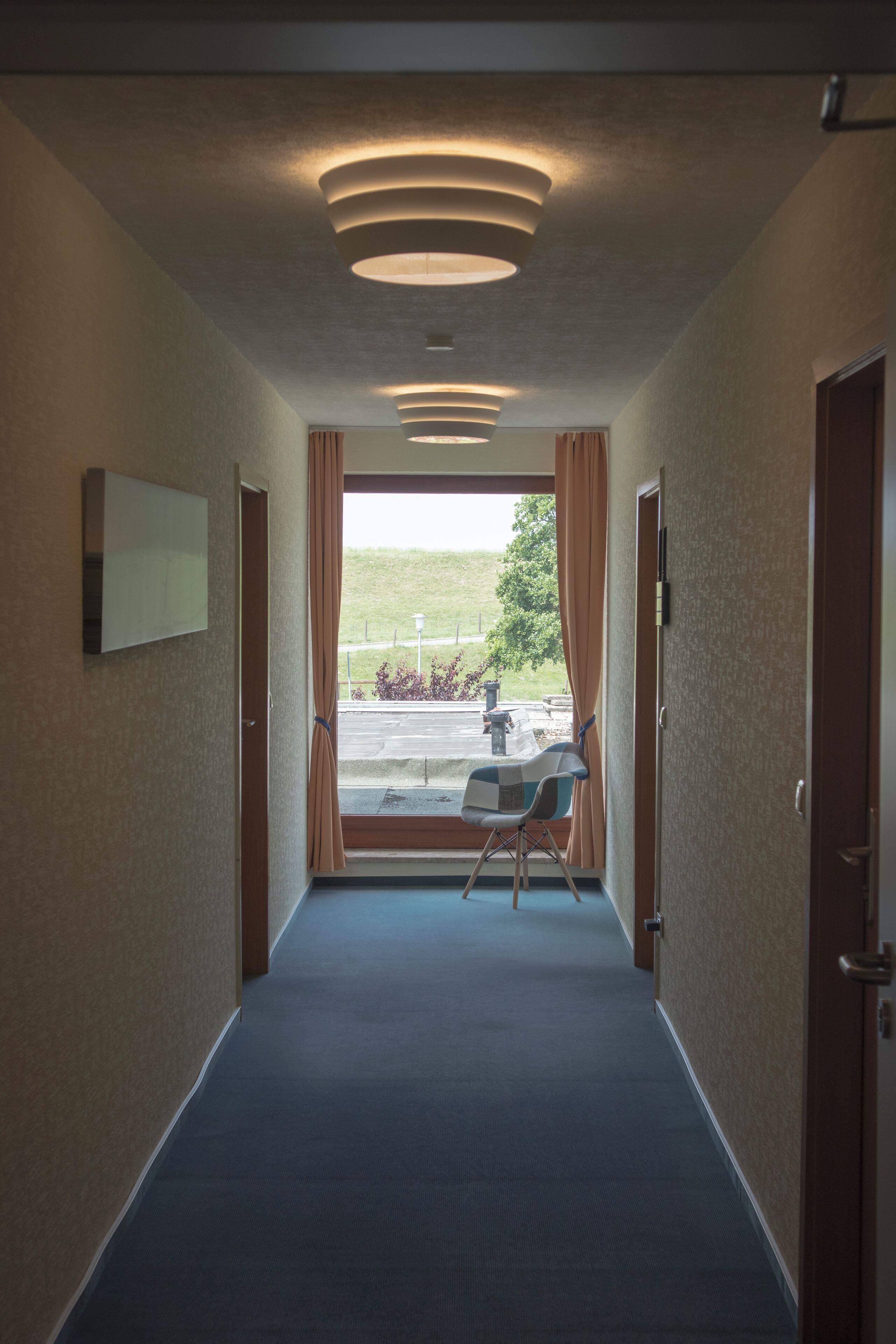 A hallway with a blue carpet, a chair, and a window with curtains.