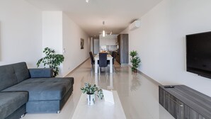 Una sala de estar moderna con un sofá, un comedor con sillas, una cocina al fondo y un televisor montado en la pared.