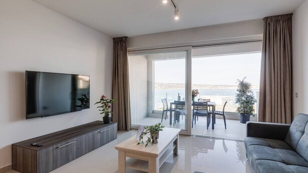Una sala de estar moderna con un televisor de pantalla plana, una consola de madera, una mesa de centro y un sofá gris. Hay un balcón con una mesa y sillas, y vistas al mar.