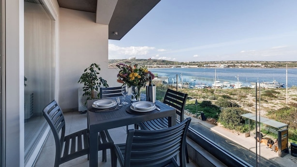 A balcony with a table set for two, overlooking a coastal area with boats and buildings.