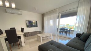 A modern living room with a dining area, a flat-screen TV mounted on the wall, and a large sliding glass door leading to an outdoor patio.