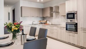 A modern kitchen with light wood cabinets, a stainless steel oven, and a microwave. There is a dining area with a table set for two and a vase of flowers.