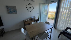 Une pièce avec une table en bois, des chaises blanches, une horloge, un bureau avec un ordinateur portable, une plante et une vue à travers une porte vitrée.