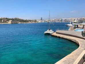 A marina with boats docked, a clear blue sea, and buildings in the background.