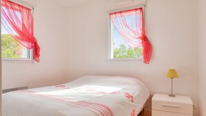 A bedroom with a bed, a nightstand with a lamp, and two windows with sheer curtains. - Close to beaches and shops, flat for 4 (Fouesnant)