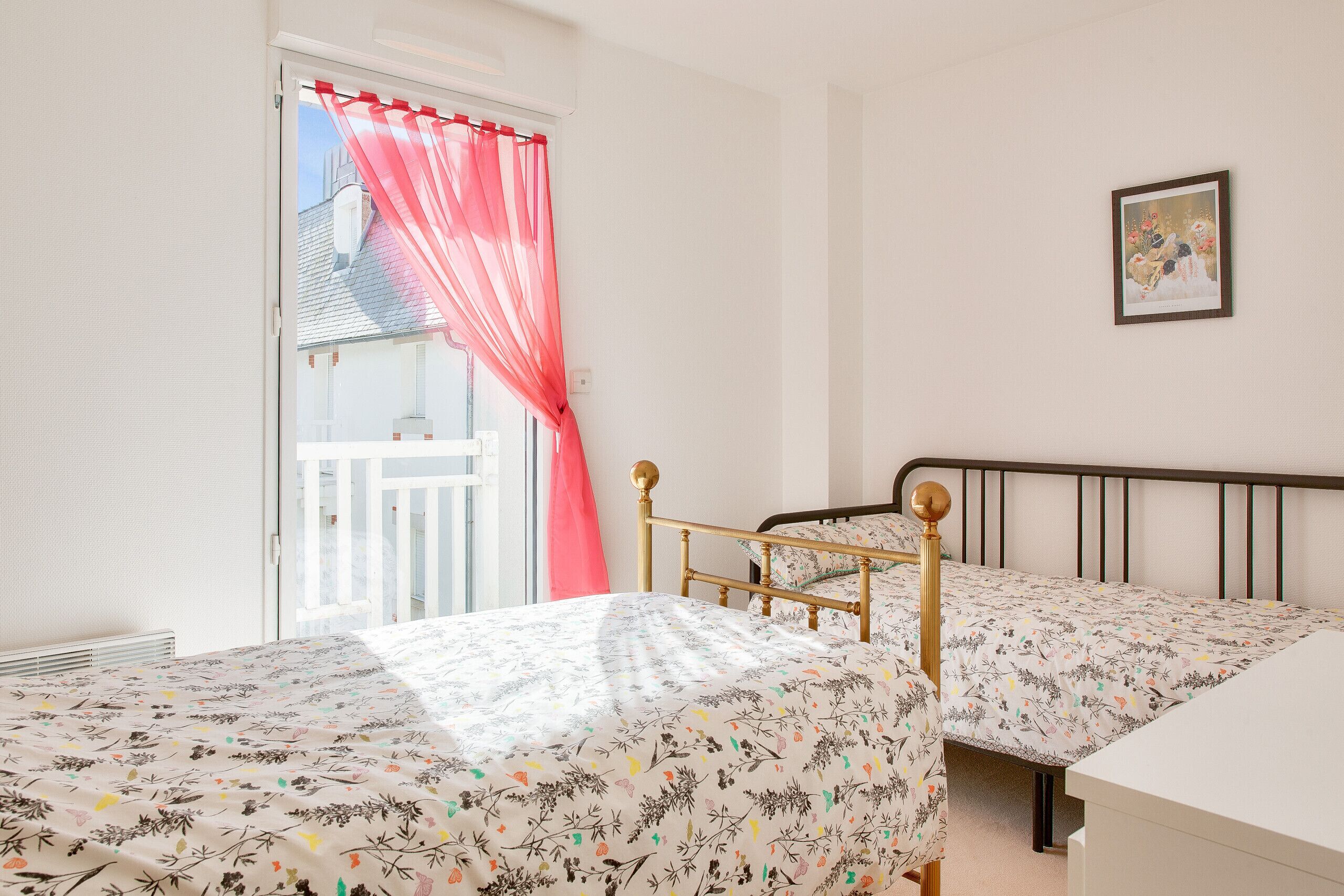 Un dormitorio con dos camas, un balcón con cortina roja y un cuadro enmarcado en la pared.