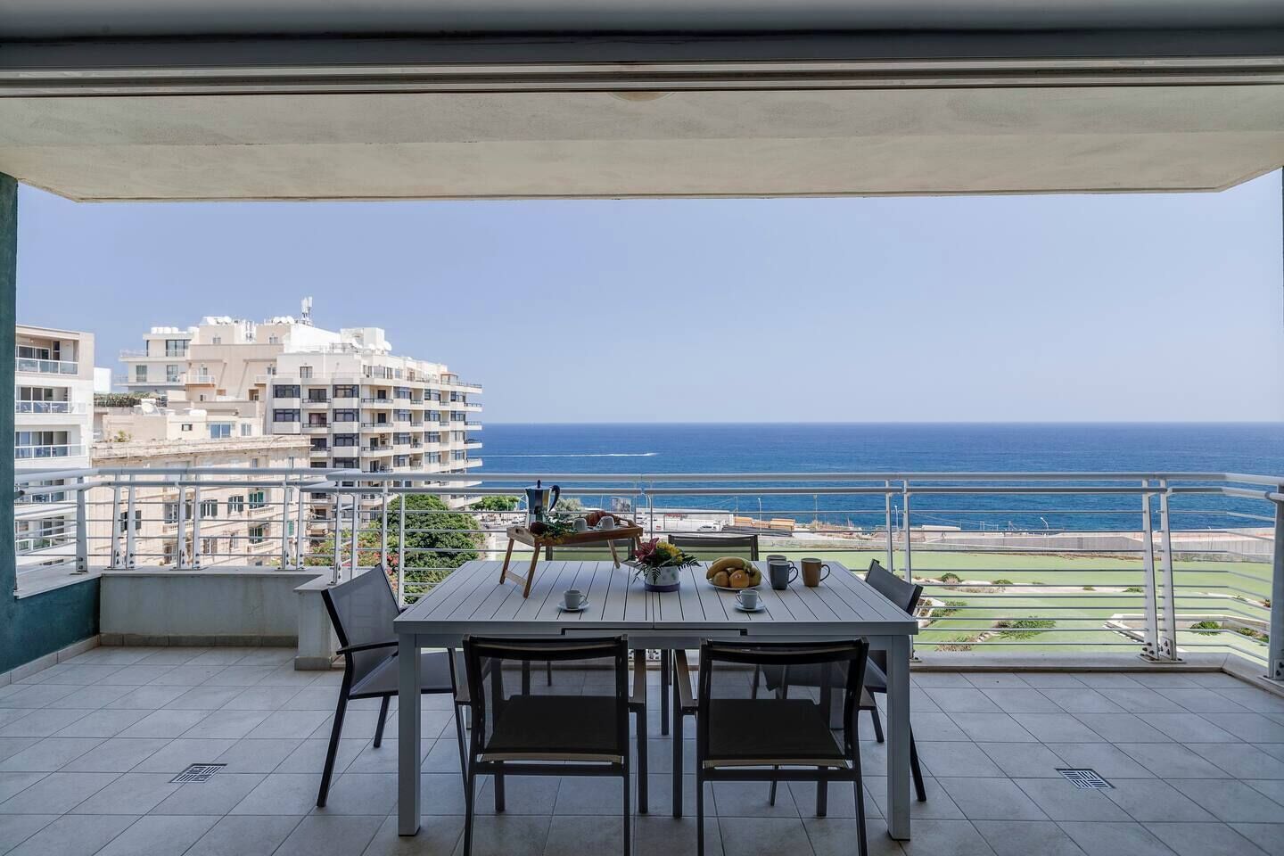 Un balcon avec une table dressée pour deux personnes, offrant une vue sur la côte, avec des bâtiments et la mer en toile de fond.