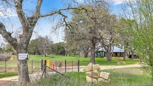 A fenced entrance to Green House with a sign, a path leading to a building, and a large rock formation. - Expansive Hill Country Ranch w/ Pool Access on 2,001 Acres (Johnson City)