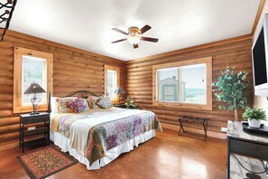 A log cabin bedroom with a bed, two nightstands, a ceiling fan, and a large window. - Lighthouse Hill Ranch - Log Home - situated on 2001 acres! (Johnson City)