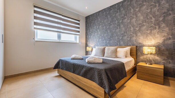 A modern bedroom with a large bed, a nightstand with a lamp, and a window with blinds.