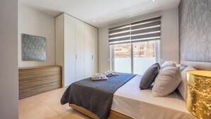 A modern bedroom with a bed, a window with blinds, a headboard, a bedside table with a lamp, and a wall-mounted artwork.