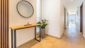 Un pasillo moderno con suelo de madera, un espejo redondo, una mesita con una planta y vistas al mar a través de una puerta abierta.