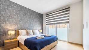 Un dormitorio moderno con una cama grande, una mesita de noche con lámpara y vistas a través de una ventana con persianas.