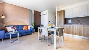 A modern living room with a blue sofa, a dining table with chairs, and a kitchen area with white cabinets and a wooden accent wall. - Family-friendly apartment near Ghadira, Paradise & Golden Bay Modern amenities & coin-operated A/C Perfect base for your Malta beach getaway. (Il-Mellieħa)