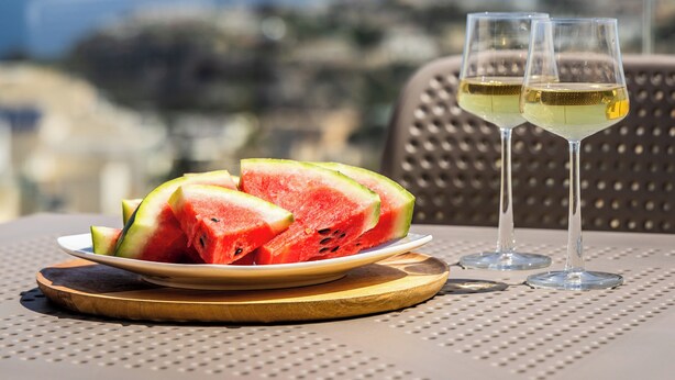 A table with a plate of watermelon slices and two glasses of white wine, set against a cityscape view. - Family-friendly apartment near Ghadira, Paradise & Golden Bay Modern amenities & coin-operated A/C Perfect base for your Malta beach getaway. (Il-Mellieħa)