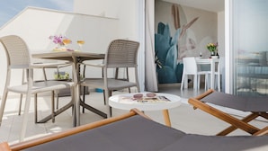 A balcony with a table set for two, two chairs, a lounge chair, and a glass door leading to a room with a large plant.