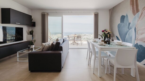 A modern living room with a sofa, dining table, and chairs, a large window with a city view, and a painting on the wall.