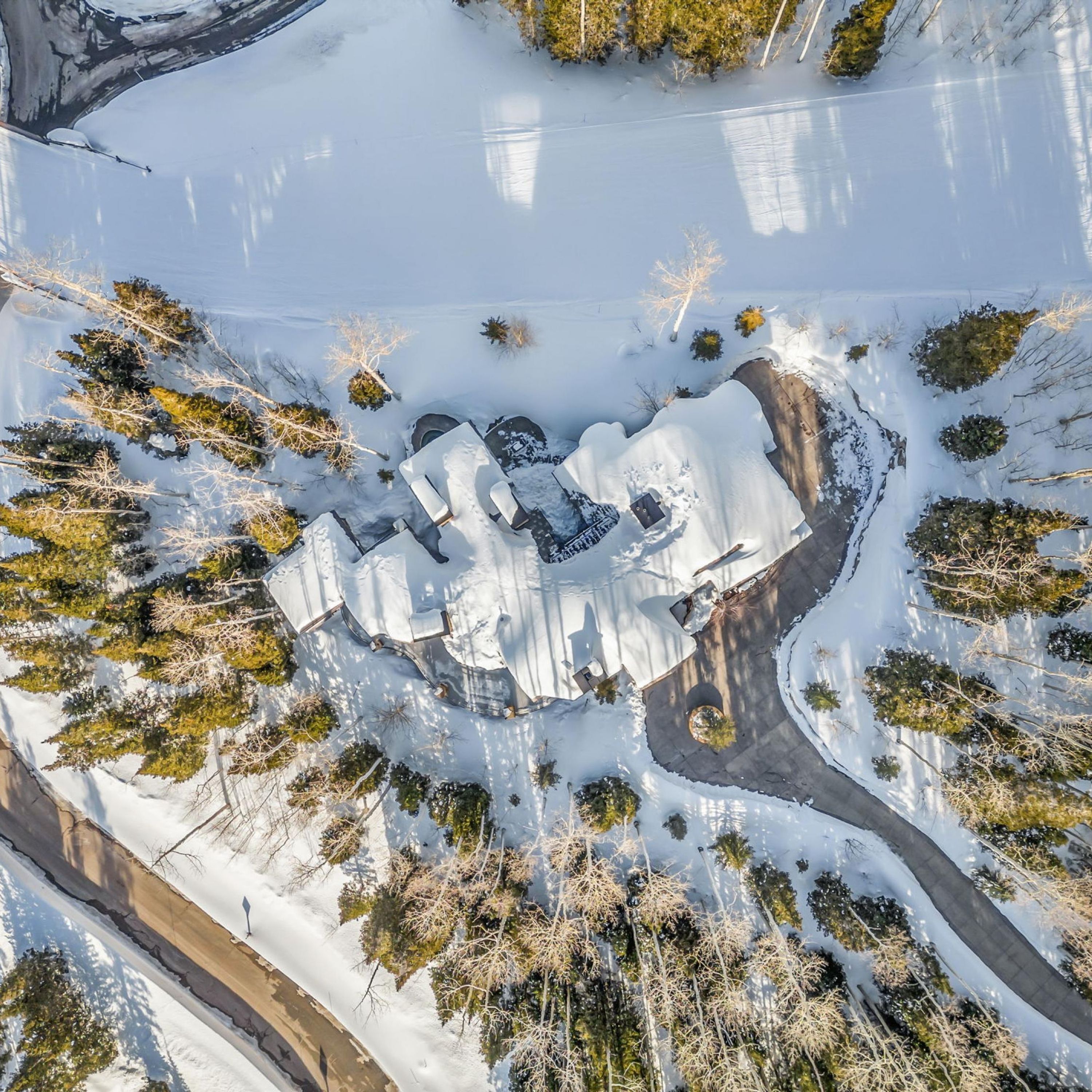 Eine Vogelperspektive zeigt eine schneebedeckte Landschaft mit einer kreisförmigen Anordnung von Gebäuden, umgeben von Bäumen und einer verschneiten Straße.