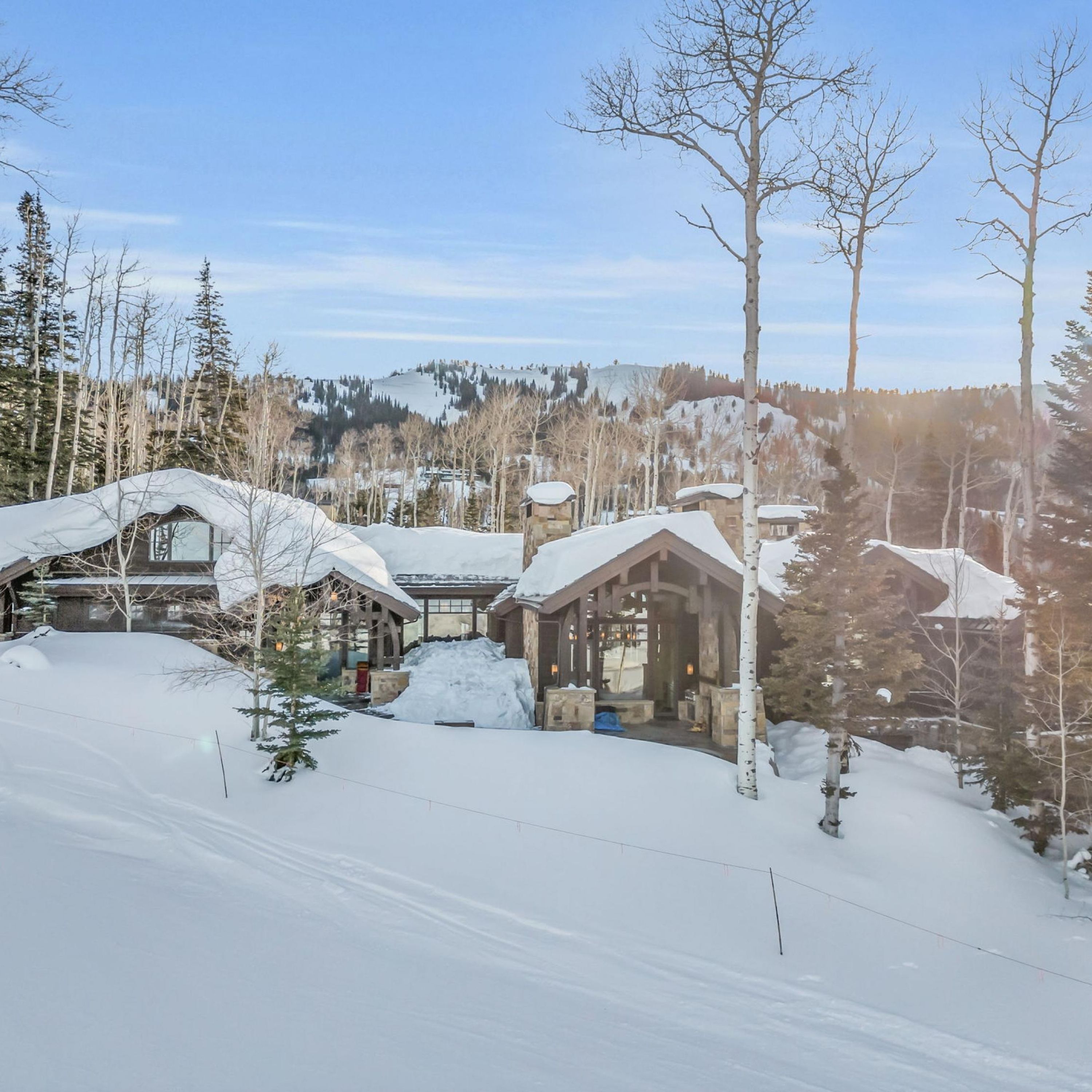 A snow-covered lodge with multiple buildings, including a main house and a smaller cabin, surrounded by trees and a mountainous backdrop.