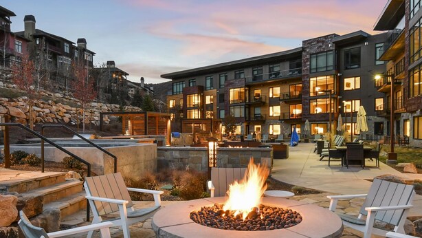 A courtyard with a stone fire pit surrounded by Adirondack chairs and a round table, overlooking a multi-story building with balconies and outdoor seating. - Lift Condo With Ski-in out w Village Access hot tub Fitness Center ski Rentals On-site (Park City)