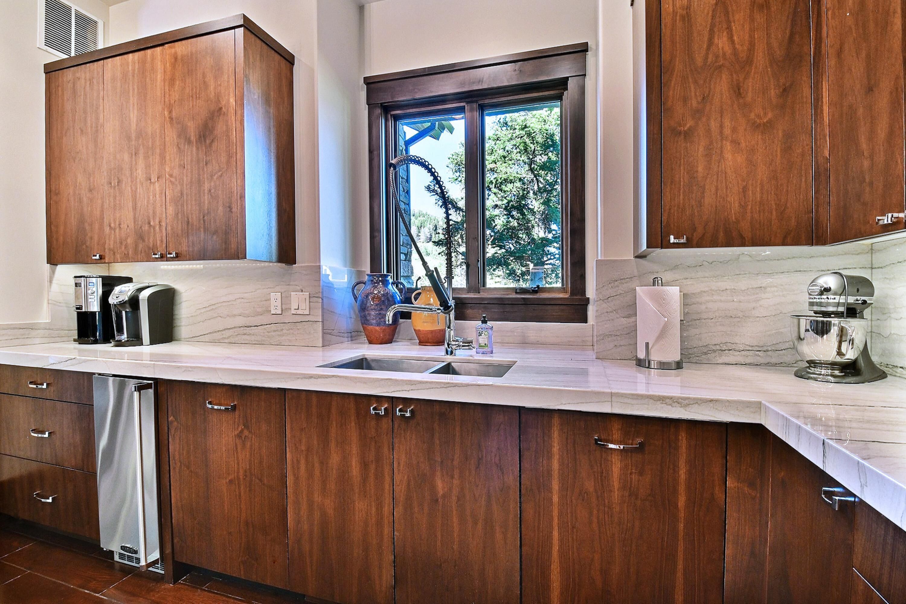 A modern kitchen with wooden cabinets, a marble countertop, a built-in dishwasher, and a window with a view of trees.