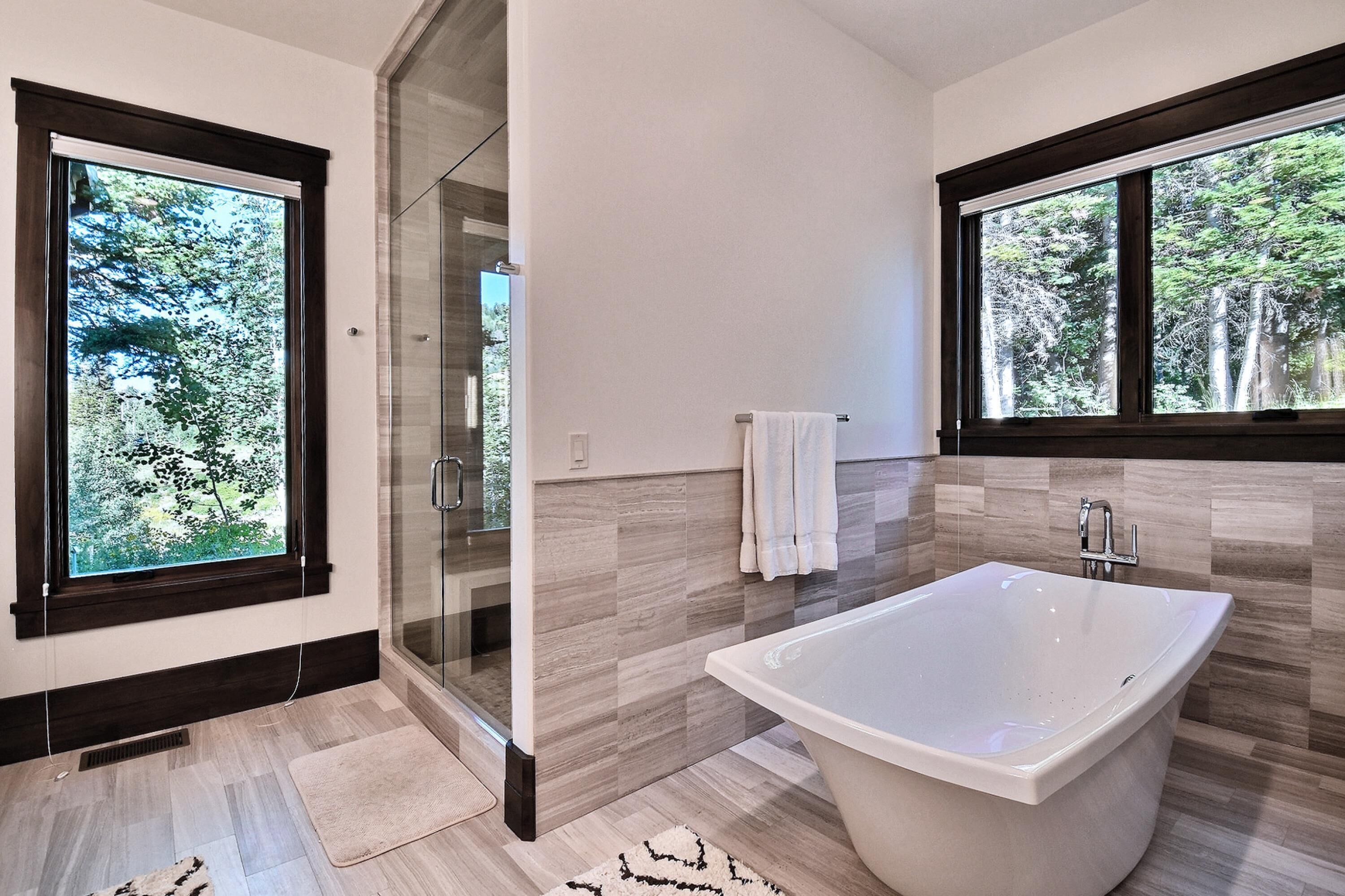 Un baño moderno con una bañera independiente, cabina de ducha de vidrio y amplias ventanas con vistas a árboles.