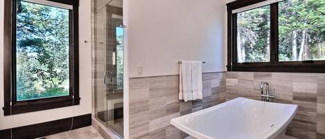Un baño moderno con una bañera independiente, cabina de ducha de vidrio y amplias ventanas con vistas a árboles.