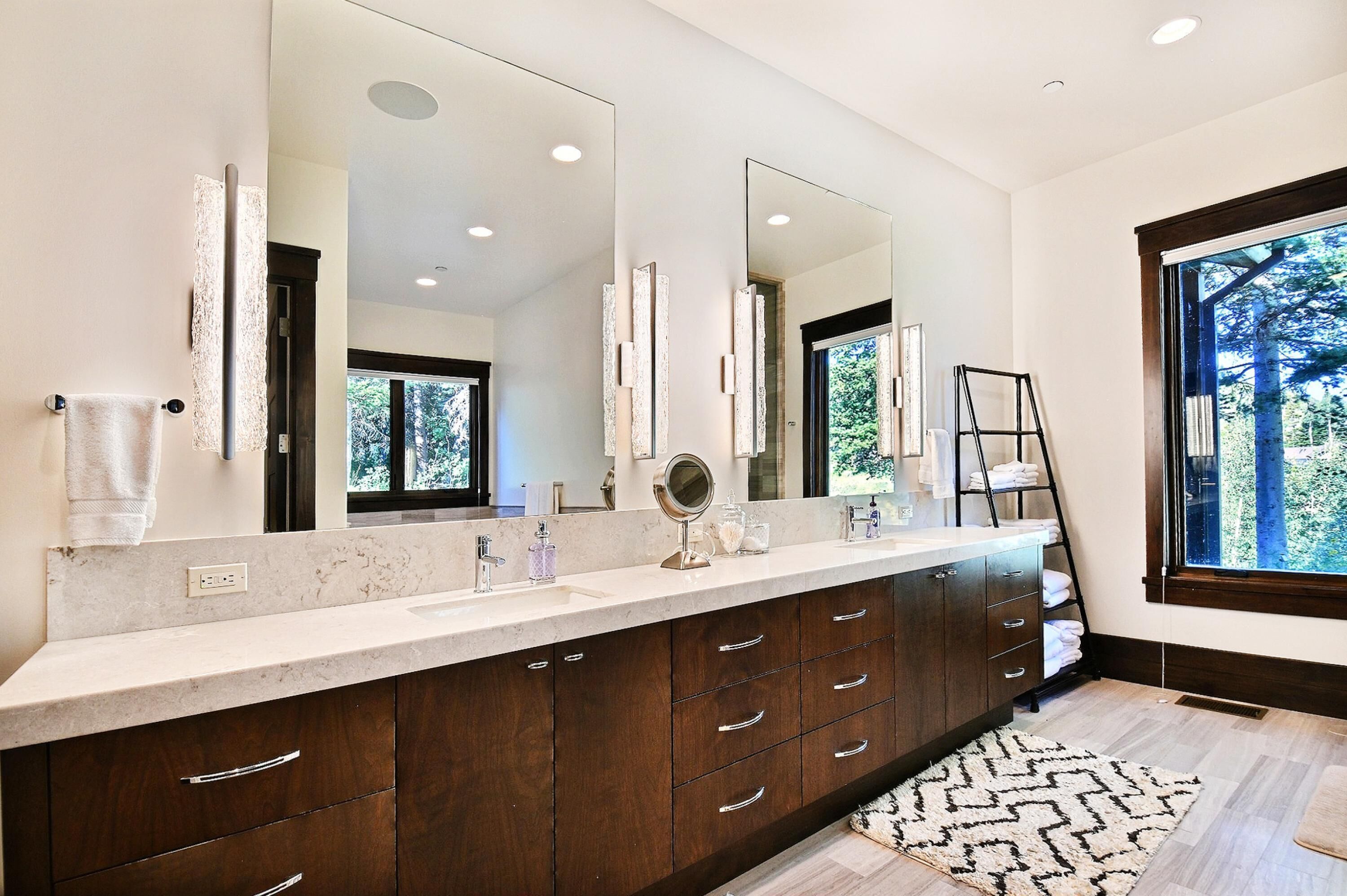 A modern bathroom with a large mirror, dark wooden cabinets, and a large window offering a view of trees.