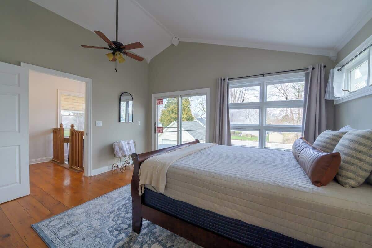 A bedroom with a large bed, a ceiling fan, a window with curtains, and a view of the outside.