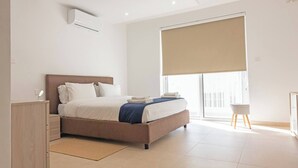 A modern bedroom with a large bed, a bedside table, a small stool, and a window with a blind.