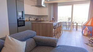 A modern apartment with a grey sofa, a kitchen with a built-in oven, a dining area with a table and chairs, and a balcony with a tent.