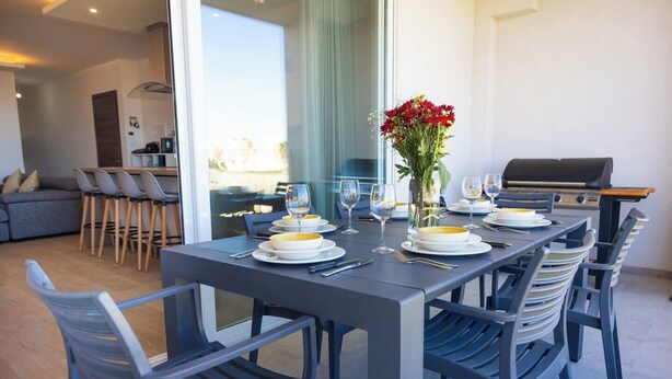 A dining area with a table set for four, chairs, a vase with flowers, and a grill in the background.