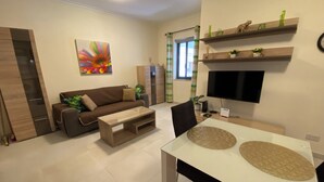 Una sala moderna con sofá, mesa de centro y televisor. Hay una mesa de comedor con sillas. La habitación tiene un ventanal con cortinas y un cuadro en la pared.