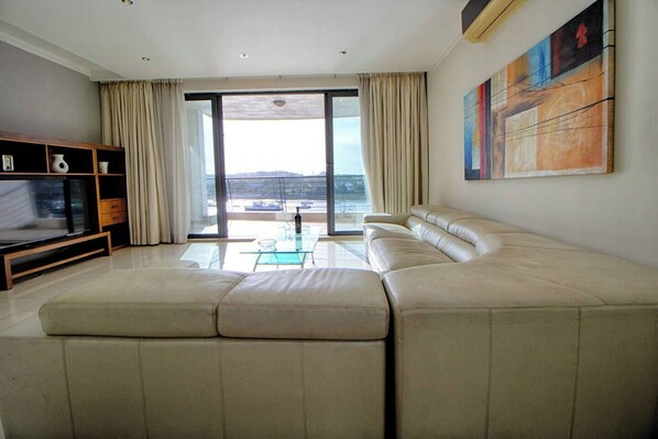 A modern living room with a large beige sectional sofa, a glass coffee table, and a TV on a wooden stand. There is a sliding glass door leading to a balcony with a view of the sea.