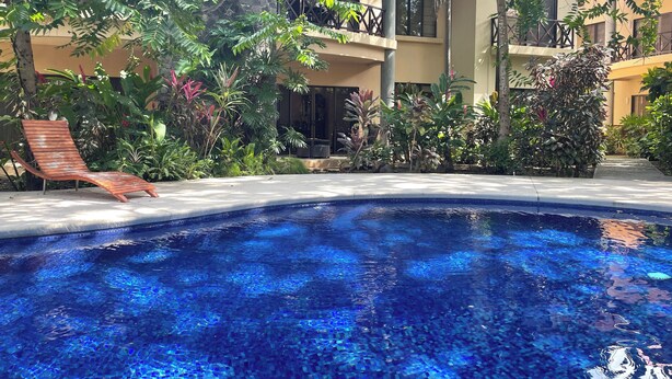 A swimming pool with clear blue water, surrounded by lush greenery and a wooden lounge chair.