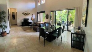 A spacious living area with a dining table, sofas, and a TV. There are windows providing natural light and a view of greenery outside.