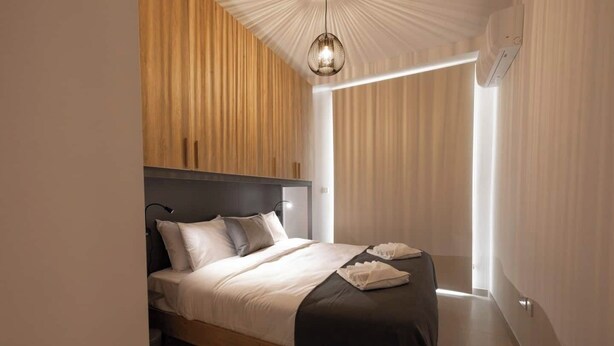A modern bedroom with a wooden headboard, a bed with white and gray bedding, a bedside lamp, and a window with sheer curtains. - Modern 2-bedroom apartment in central Mellieħa. Sleeps 6 with sofa bed, A/C, Wi-Fi, Smart TV & lift access. Steps from shops, restaurants & public transport. (Il-Mellieħa)