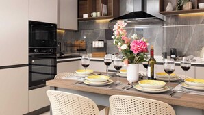 A modern kitchen with a dining area, featuring a built-in oven, a stainless steel range hood, and a wooden dining table set with plates, glasses, and a vase of flowers. - Modern 2-bedroom apartment in central Mellieħa. Sleeps 6 with sofa bed, A/C, Wi-Fi, Smart TV & lift access. Steps from shops, restaurants & public transport. (Il-Mellieħa)